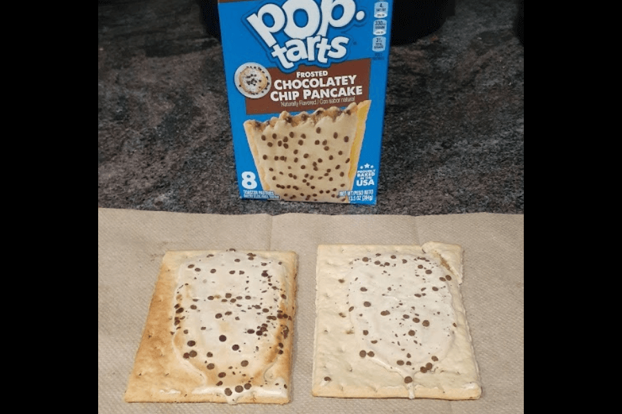 Two Frosted Chocolatey Chip Pancake Pop-Tarts are displayed on a napkin, featuring a light beige frosting with mini chocolate chips sprinkled on top. Behind them is the product box showing an image of the Pop-Tart and branding in blue and brown tones.