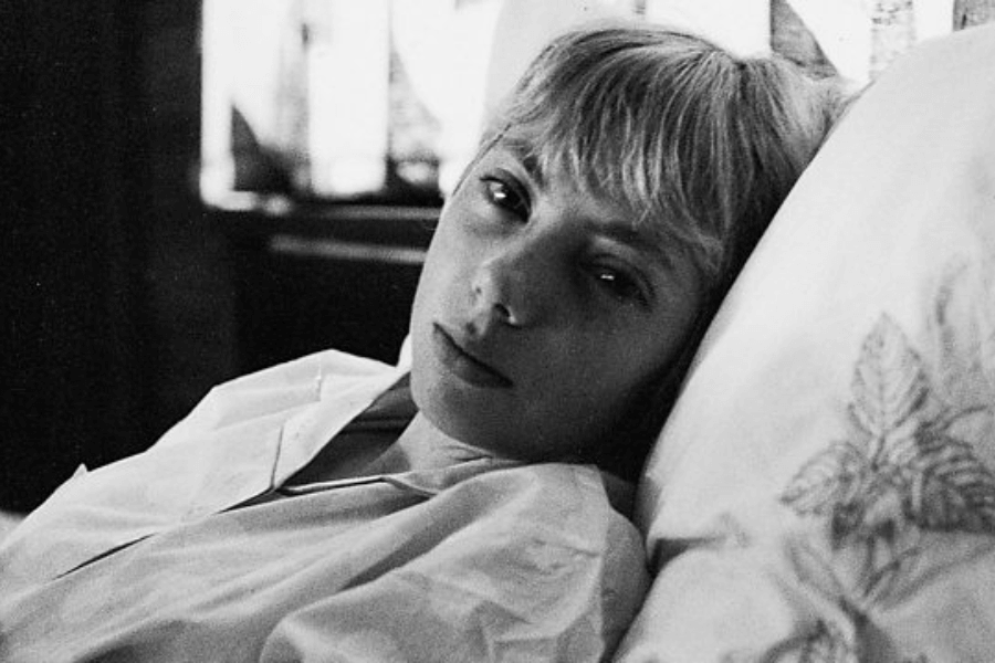 Black-and-white close-up of a young woman lying in bed, looking directly at the camera with a calm, slightly solemn expression. She is wearing a collared shirt, and a pillow with a leaf design is visible beside her.