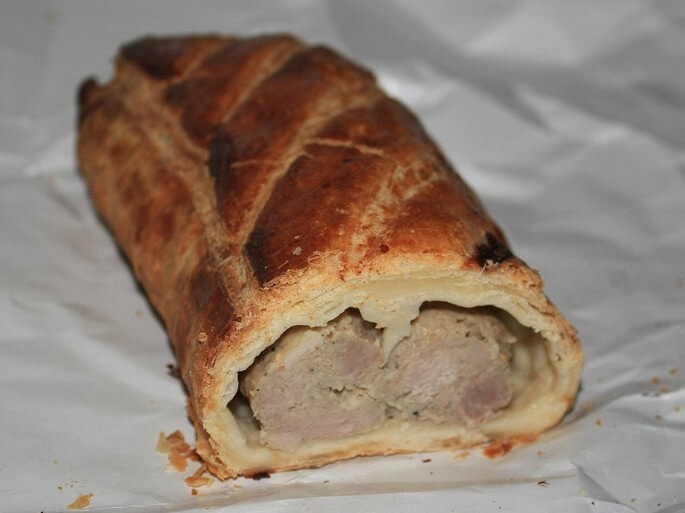 A golden-brown pastry roll sliced open to reveal a filling of sausage meat. The flaky, buttery crust has a crosshatch pattern, and some crumbs are scattered on the white paper underneath.
