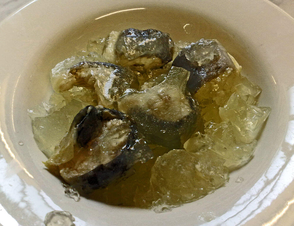 A close-up of a white plate filled with chunks of fish encased in a translucent yellowish aspic. The gelatin surrounds the fish pieces, creating a shiny, wobbly texture, with light reflecting off the glossy surface.