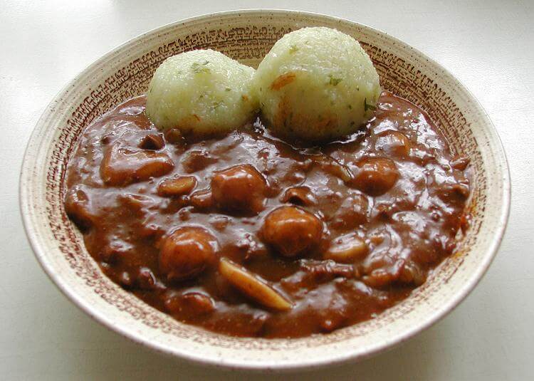 A ceramic bowl filled with a thick, dark brown stew containing small sausages and sliced mushrooms. Two round, fluffy potato dumplings sit on top of the stew, slightly browned and garnished with herbs.