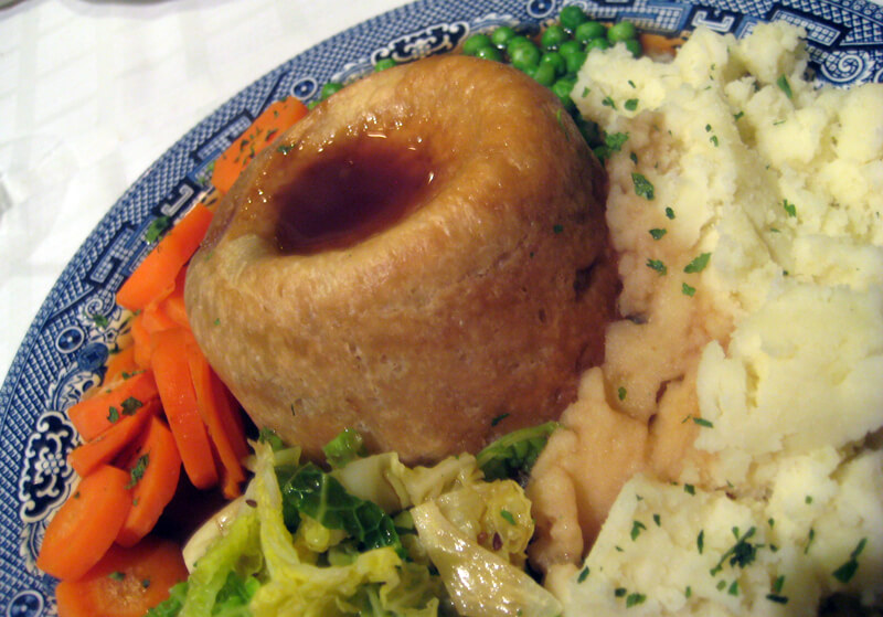 A plate of British-style comfort food featuring a golden-brown Yorkshire pudding filled with gravy. Surrounding it are mashed potatoes, sliced carrots, green peas, and some wilted cabbage, all garnished with chopped herbs.