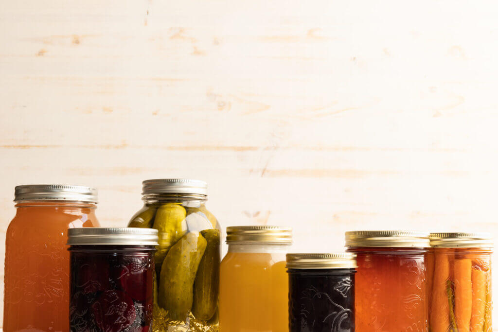 Border of glass jars, pickled beets, pickles, filled carrots, honey and apple jelly, in autumn colours on a white wood background with copy space