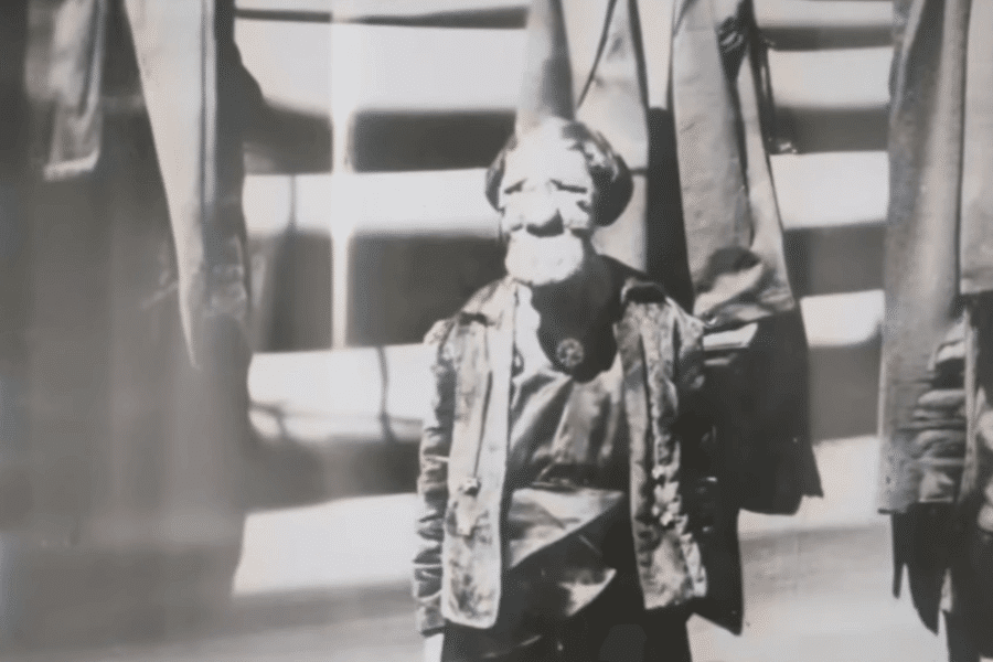 Vintage black and white photo of a person with dwarfism and a visibly disfigured face, standing in front of a staircase flanked by coats or garments hanging on either side. The person wears formal or performance-like attire, possibly suggesting involvement in public exhibition or a sideshow, reflecting a time when individuals with visible differences were often objectified for entertainment.