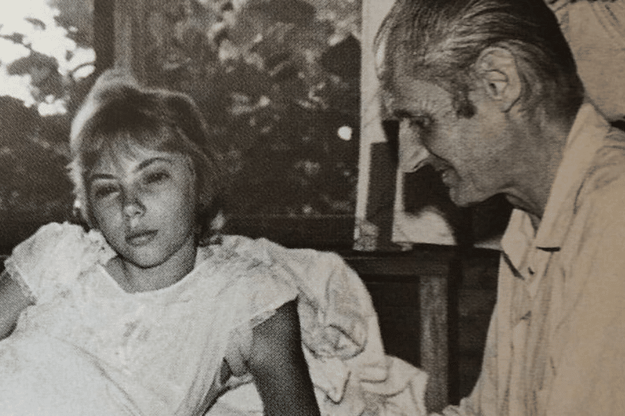 Black-and-white photo of a young woman lying in bed, appearing tired or recovering, while an older man with slicked-back hair sits beside her, smiling gently. Light filters through a window behind them, revealing blurred outdoor foliage.