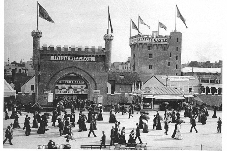 Black and white photograph of a grand entrance labeled "Irish Village" with castle-like towers and flags, part of a World's Fair or exposition. A large sign also marks “Blarney Castle” in the background. Crowds of men and women in Edwardian dress, many with parasols, stroll in front of the display, capturing a scene of cultural exhibition and early 20th-century public entertainment.