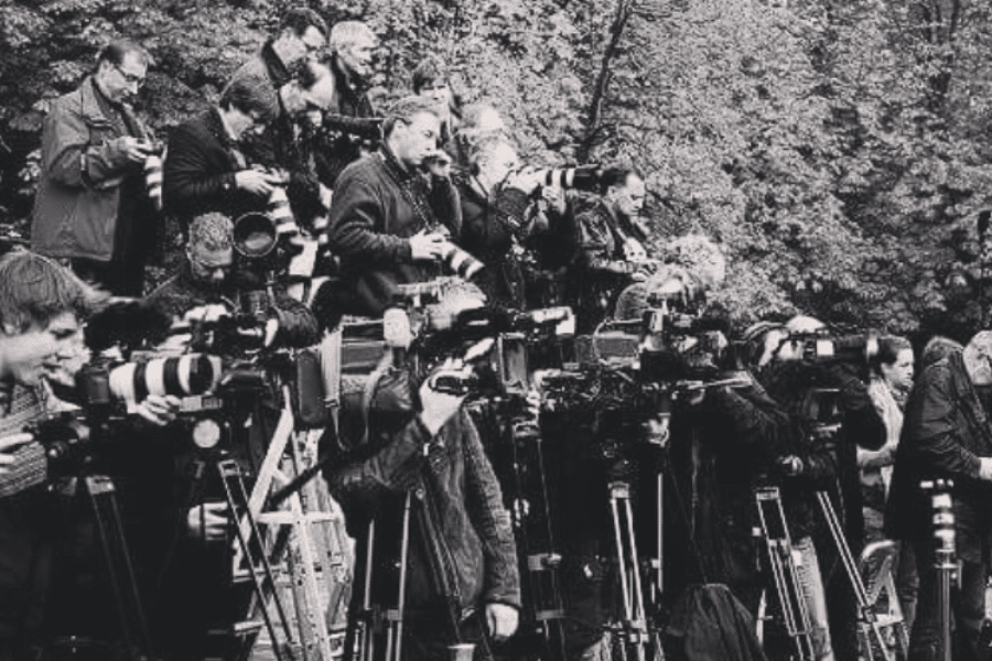 Black-and-white photo showing a large crowd of photographers and videographers, all equipped with professional cameras and long lenses, closely packed together on risers and ladders, capturing an unseen event in a wooded outdoor setting.