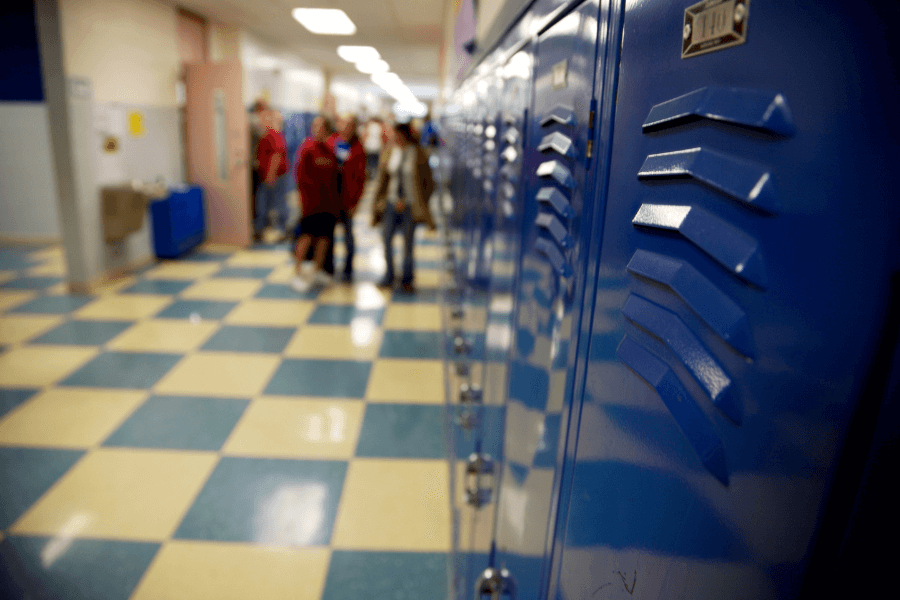 Row of shiny blue lockers in a school hallway with a checkered blue and cream floor. A group of students is blurred in the background, adding a sense of movement and daily school activity.