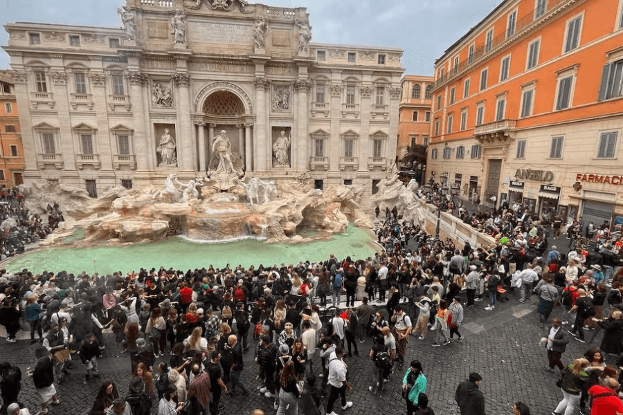 Large crowd of tourists gathered around the Trevi Fountain in Rome, admiring the grand baroque architecture and sculptures as turquoise water flows into the basin; the surrounding buildings and shops frame the bustling scene.