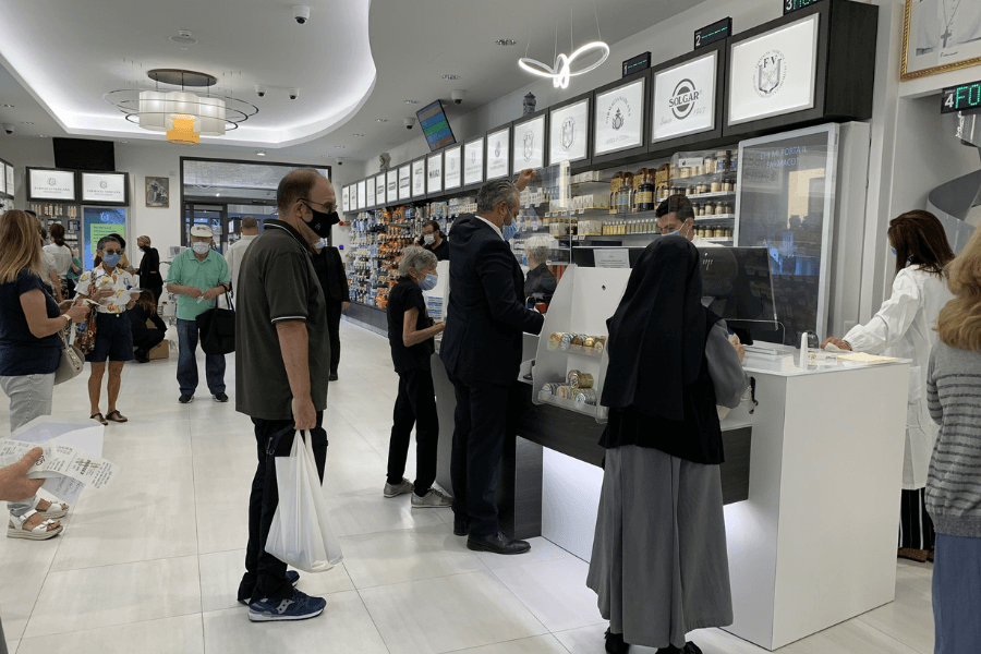 Interior of the Vatican Pharmacy, where people, including a nun and several masked customers, wait in line or interact with pharmacists behind a sleek counter; shelves are stocked with health and wellness products beneath illuminated signage.