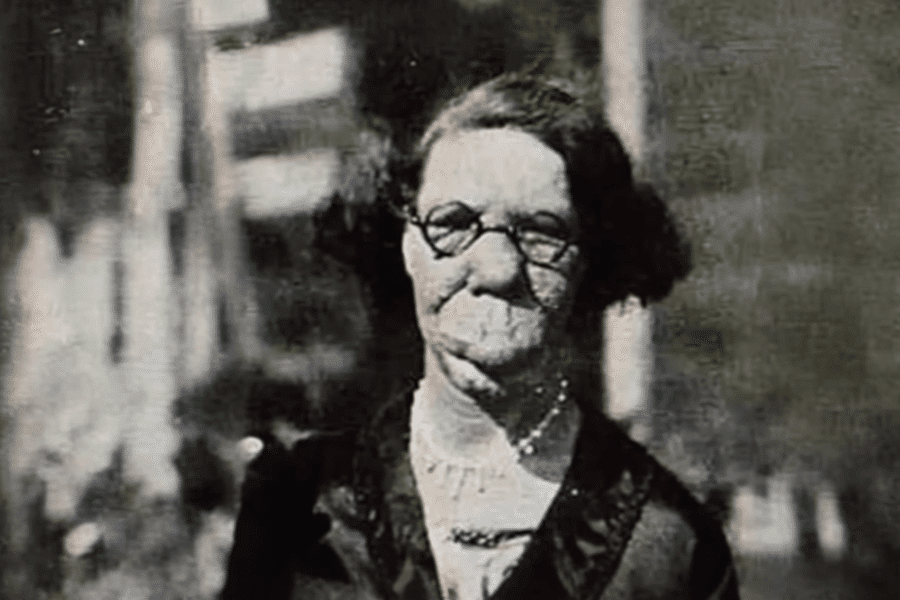 Colorized historical photo of a woman with severe facial disfigurement, likely due to a condition such as neurofibromatosis or another congenital disorder. She wears round glasses, a dark dress with a white collar, and a necklace, and gazes calmly at the camera. The background is softly blurred, emphasizing her presence and dignity in the face of a world that often marginalized visible difference.