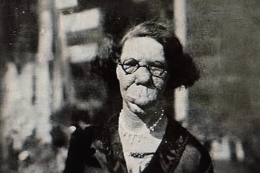 Colorized historical photo of a woman with severe facial disfigurement, likely due to a condition such as neurofibromatosis or another congenital disorder. She wears round glasses, a dark dress with a white collar, and a necklace, and gazes calmly at the camera. The background is softly blurred, emphasizing her presence and dignity in the face of a world that often marginalized visible difference.