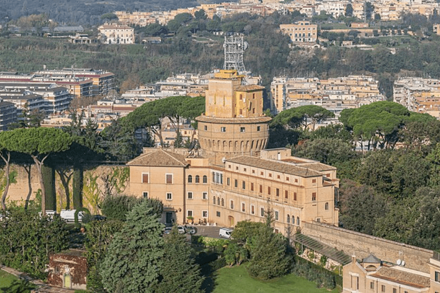 Aerial view of the Vatican Radio building surrounded by lush gardens and stone walls, with a central cylindrical tower topped with antennas and broadcasting equipment, set against a backdrop of modern buildings and green hills.