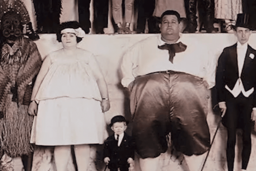 Colorized vintage photograph of a group of sideshow performers posed for a publicity photo. Among them are a man with extreme obesity, a woman labeled as a “fat lady,” a little person in formalwear, a man in a tribal costume and mask, and a tall thin man in a tuxedo. The image reflects the exploitative "freak show" era, when individuals with physical differences or non-Western heritage were exhibited for public amusement.