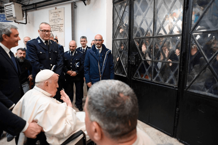 Pope Francis, seated in a wheelchair, gestures while visiting a prison facility, surrounded by guards and officials, as inmates press their hands against a barred glass door to watch him from behind.