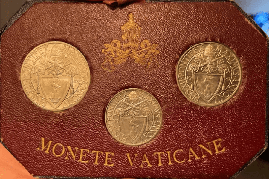 Close-up of three Vatican coins mounted on a red leather display with gold embossed text reading "MONETE VATICANE," featuring intricate papal emblems including the crossed keys and tiara, along with the Latin inscription "PIVS XII PONTIFEX MAXIMVS."