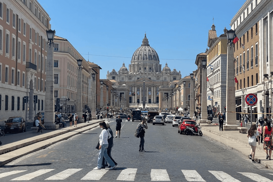 Street view from Via della Conciliazione leading up to St. Peter’s Basilica in Vatican City, with tourists crossing the cobblestone road and cars parked along the sides, framed by historic buildings and obelisks under a clear blue sky.