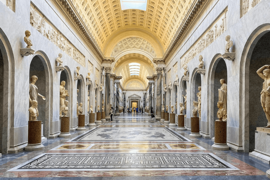 Interior view of the Gallery of the Candelabra in the Vatican Museums, featuring a long marble corridor lined with classical Roman statues and busts in arched niches, intricate mosaic flooring, and an ornately coffered ceiling illuminated by natural light from skylights.