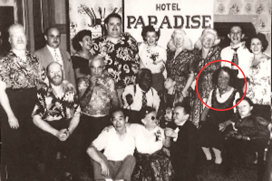 Black and white group photo of sideshow performers and entertainers gathered under a “Hotel Paradise” banner, many smiling and dressed in patterned clothing. The woman circled on the right is Grace McDaniels, identifiable by her facial disfigurement associated with Sturge-Weber syndrome. This candid moment reflects solidarity and shared life among performers who often lived on the fringes of mainstream society.