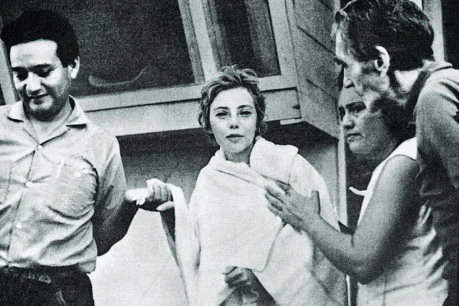 Black-and-white photo of a young woman wrapped in a towel being supported by three concerned adults, two men and one woman. She appears calm but visibly exhausted, likely in a moment of recovery or rescue, standing outside a building.