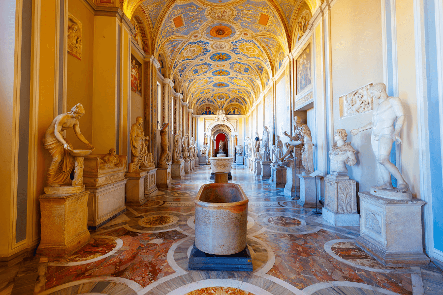 Lavishly decorated hall in the Vatican Museums, featuring a series of classical marble statues lining both sides, ornate multicolored marble floors, a central ancient bathtub, and a vaulted ceiling painted with golden and blue frescoes.