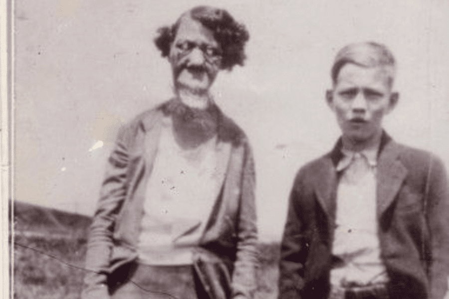 Faded vintage photo of Grace McDaniels, a woman with a severe facial deformity from Sturge-Weber syndrome, standing outdoors beside a young boy in a suit jacket and collared shirt. Both look directly at the camera with neutral expressions. The background suggests a rural or open landscape, and the image likely captures a personal moment between Grace and her son, offering a rare humanizing glimpse into their life outside of the sideshow circuit.