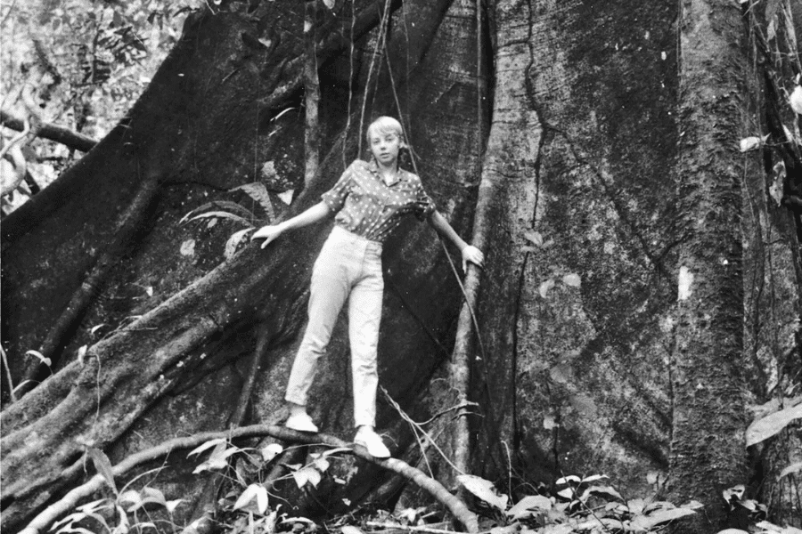 Black-and-white photo of a young woman standing on thick roots in front of a massive tree trunk in a dense jungle. She balances with one hand holding a vine, wearing a patterned shirt and light-colored pants, surrounded by lush vegetation.