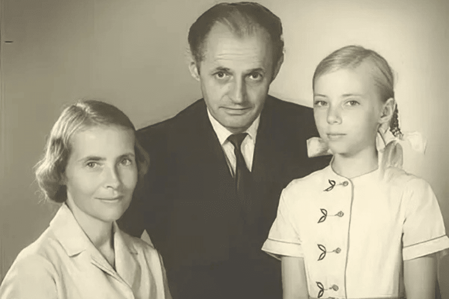 Sepia-toned studio portrait of a family, featuring a woman in a light blouse, a man in a dark suit and tie, and a young girl with braided blonde hair tied with large white bows, wearing a buttoned dress with leaf-shaped accents.