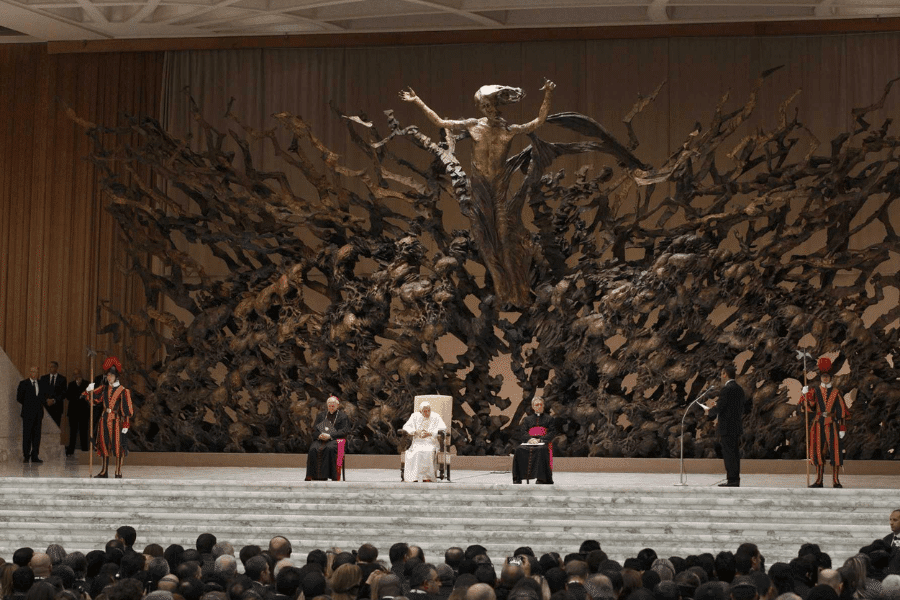 Pope presides over an audience in the Paul VI Audience Hall at the Vatican, seated in front of the dramatic bronze sculpture La Resurrezione depicting Christ rising from chaos, flanked by Swiss Guards and clergy as attendees watch from below.