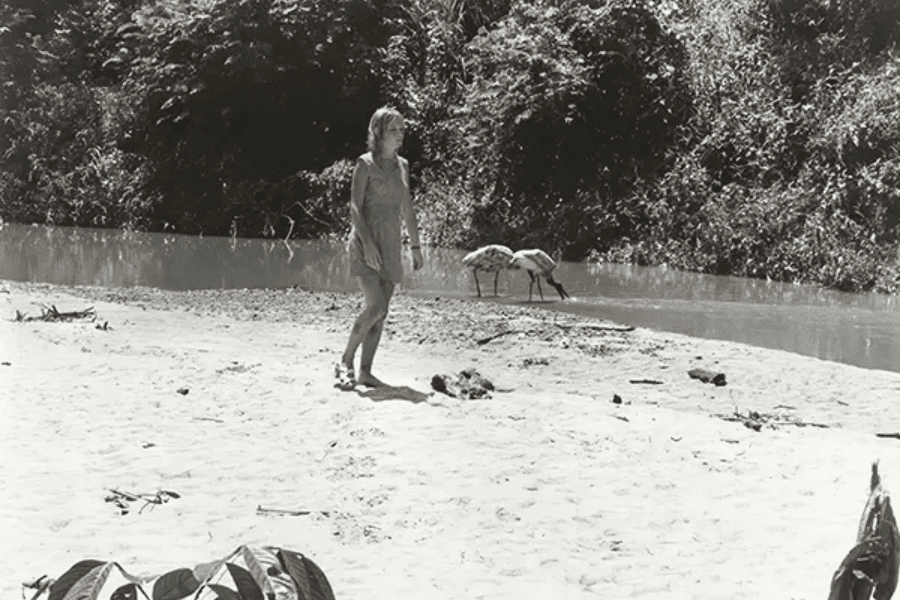 Black-and-white photo of a woman walking barefoot on a sandy riverbank, wearing a simple dress and looking toward the jungle. Two large birds are seen drinking from the river in the background, with dense forest foliage lining the opposite bank.