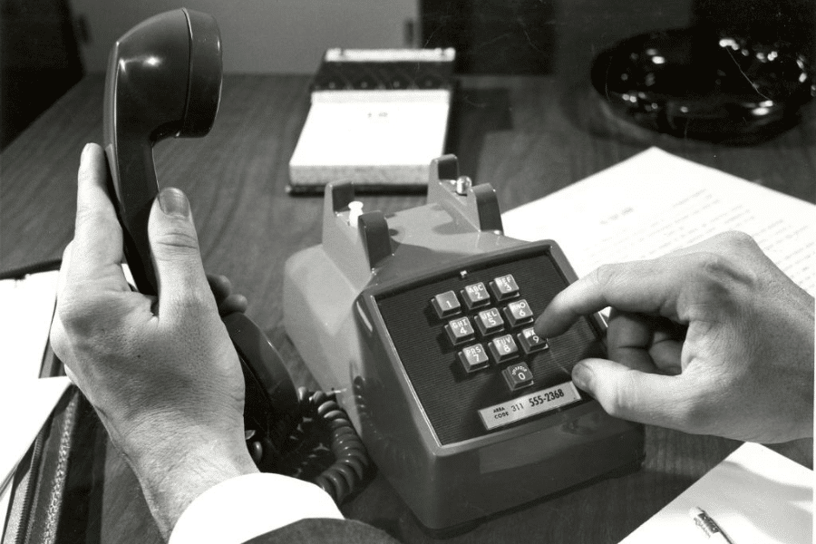 Black-and-white image of a person dialing a number on a vintage push-button desk phone while holding the receiver in the other hand, with office papers, a pen, and a calendar visible on the wooden desk.
