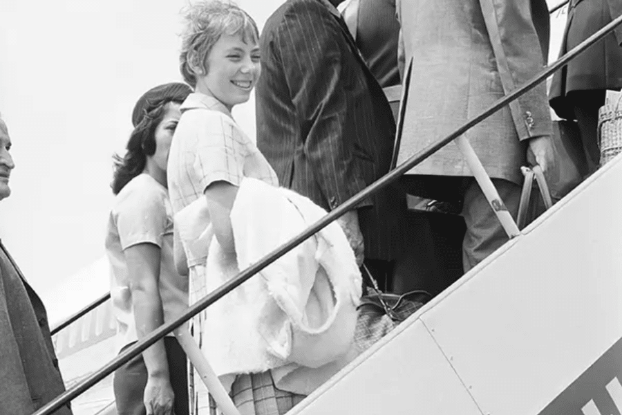 Black-and-white photo of a smiling young woman boarding an airplane via a stairway, surrounded by other passengers in formal clothing. She carries a folded blanket or coat, looking back toward the camera as she ascends.