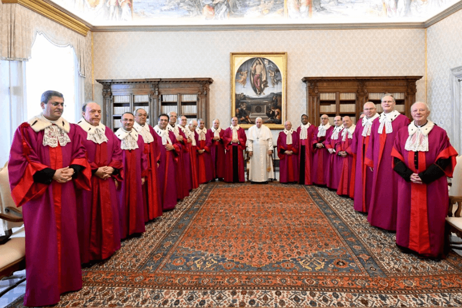 Pope Francis stands at the center of a formal group portrait with Vatican judges wearing magenta judicial robes and white lace collars, gathered in a lavish room with classical decor, wooden bookshelves, and a large religious painting in the background.