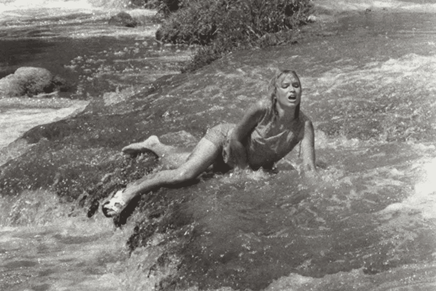 Black-and-white photo of a young woman struggling to move through a fast-flowing stream or river, visibly distressed as she braces herself against the current with one hand. Her clothing is soaked, and her body language conveys exhaustion and urgency.