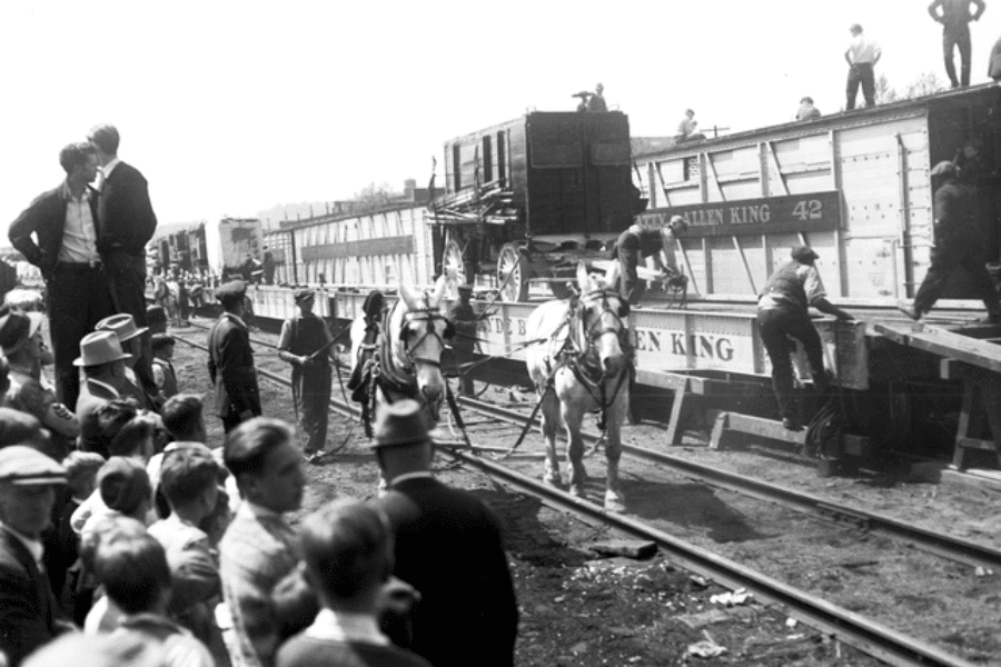 Black and white photo capturing a busy circus train unloading scene, with horses pulling ornate wagons down ramps from flatbed railcars labeled “Al G. Barnes” and “Allen King.” Workers and handlers guide the animals while crowds of onlookers, including many children, gather along the train tracks to watch. The moment reflects the behind-the-scenes labor of traveling circuses in the early 20th century and the public fascination with spectacle and mobility.