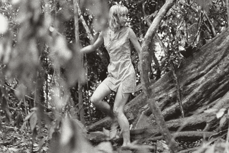 Black-and-white photo of a young woman cautiously moving through a dense jungle, barefoot and wearing a sleeveless dress. She grips vines for support while stepping over roots and uneven terrain, glancing behind her with a tense expression.