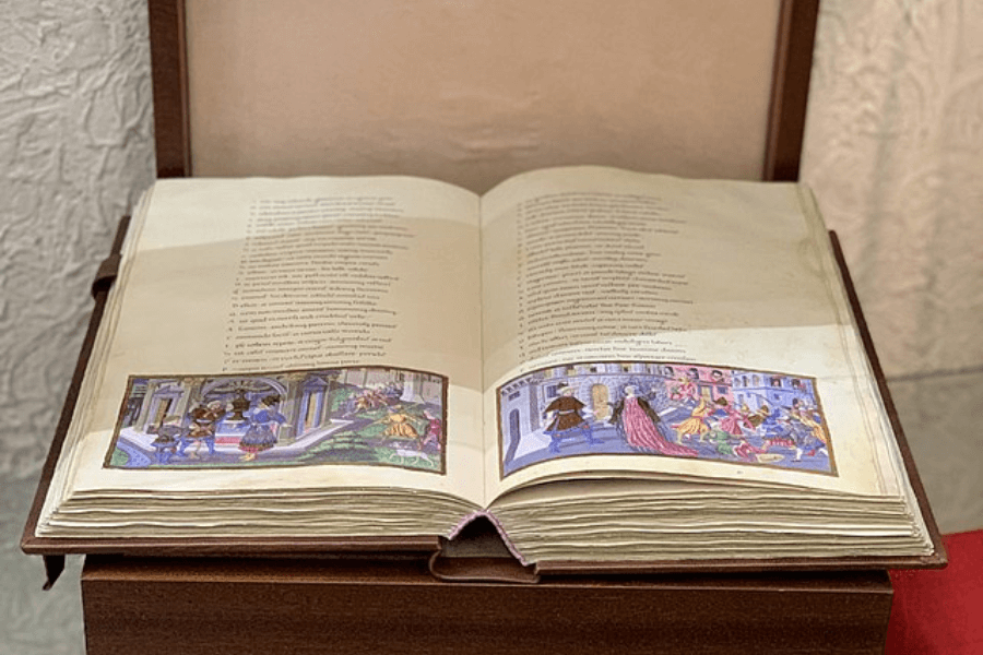 Open illuminated manuscript on display, showcasing two pages of text written in Latin with colorful miniature illustrations at the bottom depicting scenes of medieval life and architecture.