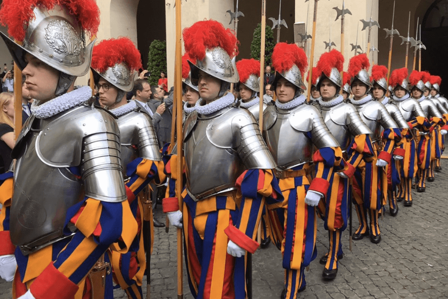 A row of Swiss Guards in traditional Renaissance-era uniforms, featuring blue, red, and yellow stripes, silver armor, and helmets topped with red plumes, marching in formation on a cobblestone path during a Vatican ceremony.