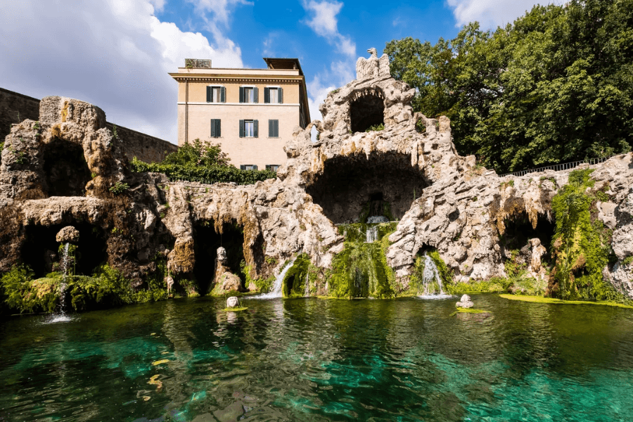 Scenic view of the Vatican Gardens featuring a naturalistic rock grotto with cascading waterfalls flowing into a crystal-clear pond, surrounded by lush greenery and with a classical building in the background under a partly cloudy sky.