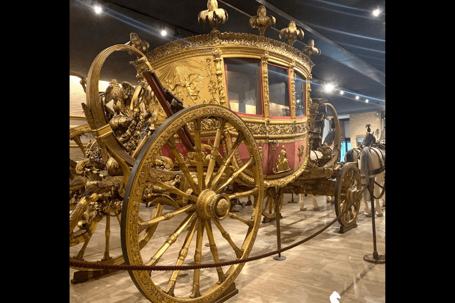 Ornate golden papal carriage on display in a museum, featuring elaborate sculptural details, red velvet interior, and gilded wheels, surrounded by a barrier rope and spotlighted under a dark ceiling.