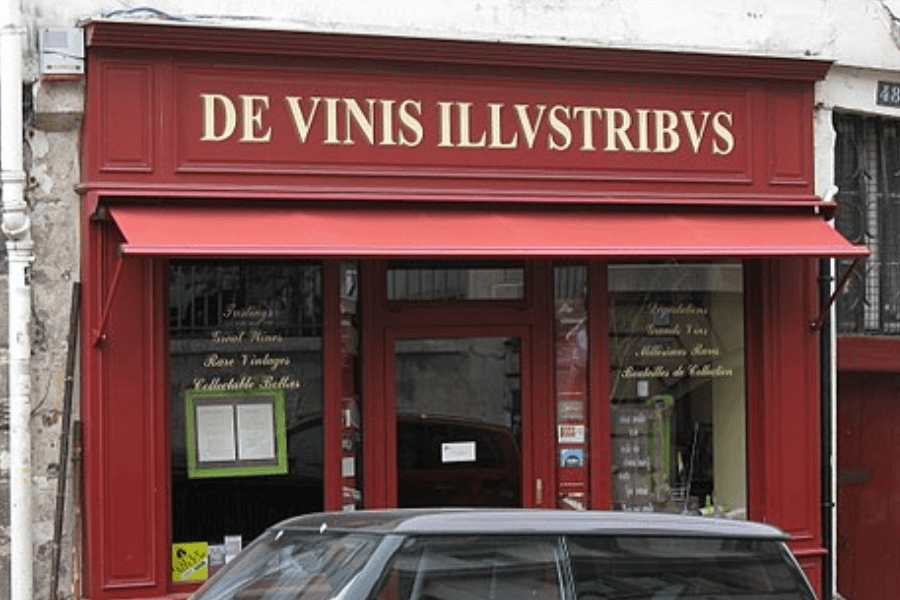 Exterior of a wine shop with a red storefront and gold lettering that reads “DE VINIS ILLVSTRIBVS,” featuring display windows advertising fine and rare vintage wines, and a parked car partially visible in front.