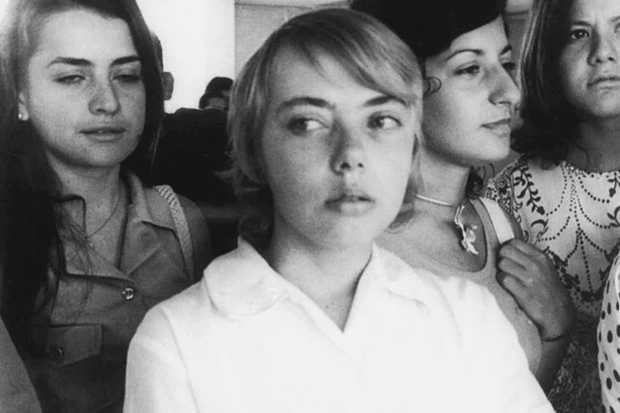 Black and white photo of a young woman in a white blouse looking off to the side, surrounded closely by other young women in a crowded indoor setting. Their expressions range from calm to contemplative, suggesting a serious or intense moment.