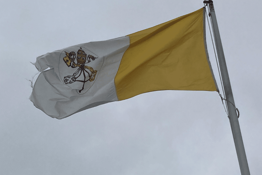 Tattered Vatican City flag flying against a gray, overcast sky, featuring two vertical bands of white and yellow with the Vatican coat of arms—consisting of the crossed keys of Saint Peter and the papal tiara—centered on the white half.