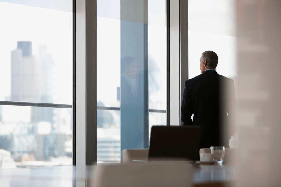 Man in a dark suit stands alone, looking out through the floor-to-ceiling windows of a high-rise office, with a view of a city skyline in the background. His reflection is visible in the glass.