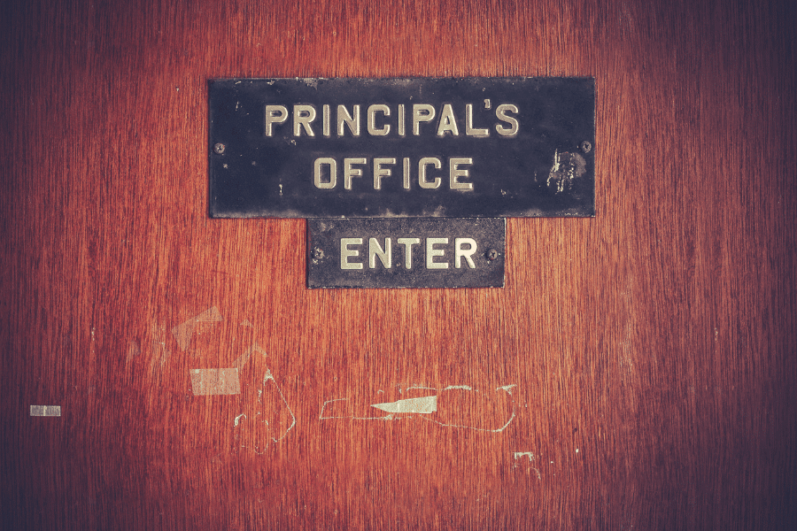 Vintage-style wooden door with a worn black sign that reads "Principal's Office" and "Enter" in bold white letters. The door has scuff marks and remnants of old tape, adding to its aged appearance.