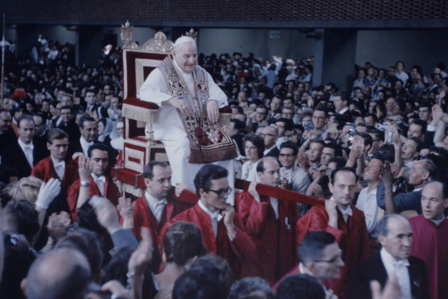 Historical photograph of Pope John XXIII being carried through a cheering crowd on the sedia gestatoria, a ceremonial throne lifted by papal attendants in red uniforms, surrounded by photographers and faithful during a Vatican event.