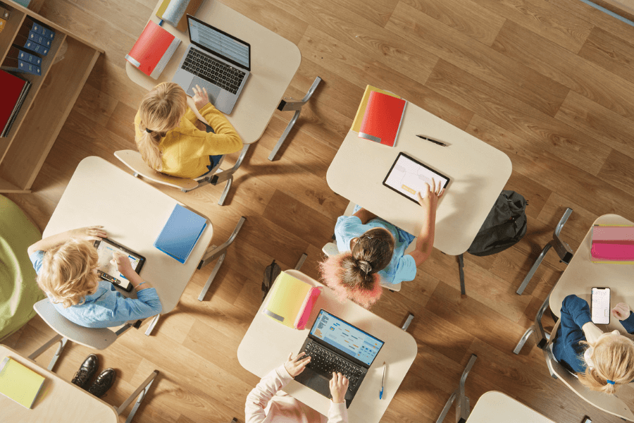 Top-down view of elementary school students sitting at individual desks in a modern classroom, each using a digital device like a tablet or laptop. The desks are organized in rows on a wooden floor, with colorful books and supplies scattered on top.