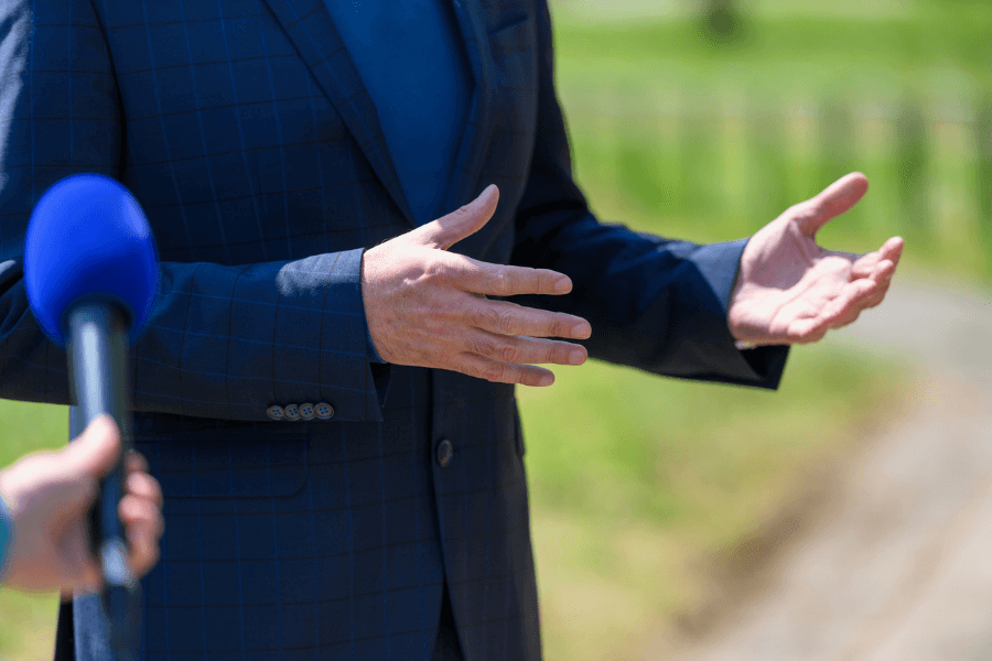 Close-up of a person in a blue checkered suit gesturing with their hands while speaking outdoors, with a blue microphone pointed toward them by someone off-camera. The background is a sunny, green outdoor setting.