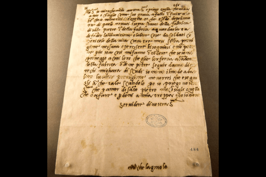 Photograph of a handwritten historical letter in Italian with brown ink on aged paper, featuring neat cursive script and signed “Michelangelo Buonarroti” at the bottom, along with an archival stamp and catalog number “484.”