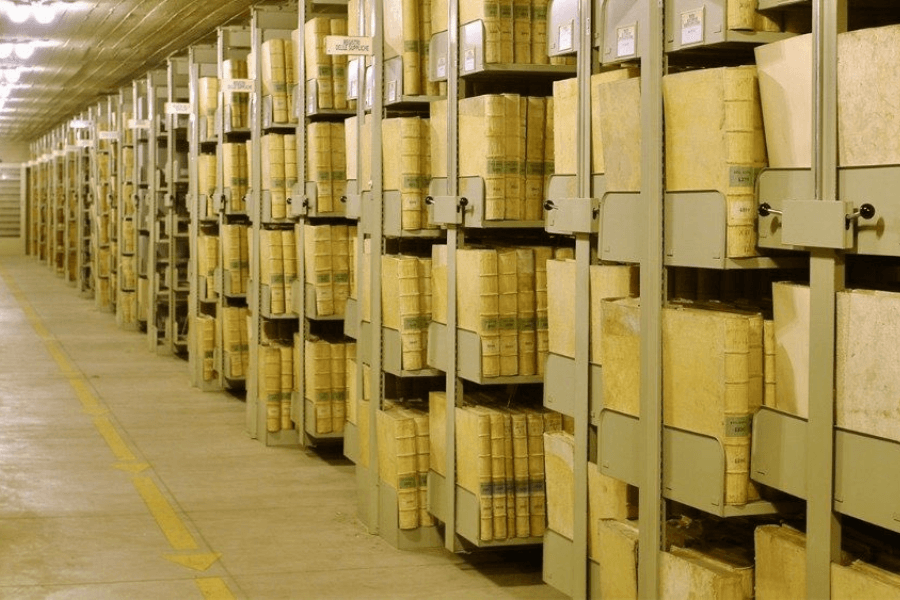Rows of metal shelving packed with large, aged volumes wrapped in parchment, stored in a climate-controlled corridor of the Vatican Apostolic Archive, formerly known as the Vatican Secret Archives.